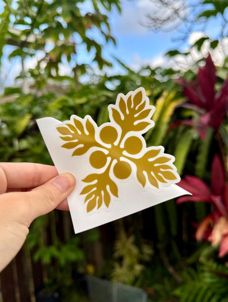 May include: A gold-colored vinyl sticker with a stylized flower design. The sticker is on a white background and is being held by a hand.