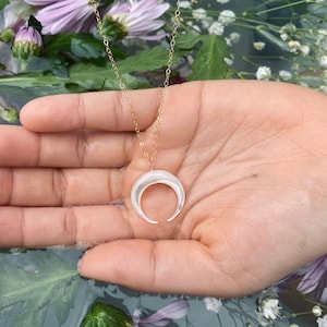 以下が含まれることがあります： 白い三日月型のペンダントが付いた金のチェーンネックレス。ネックレスは花柄の背景に置かれた手に置かれています。