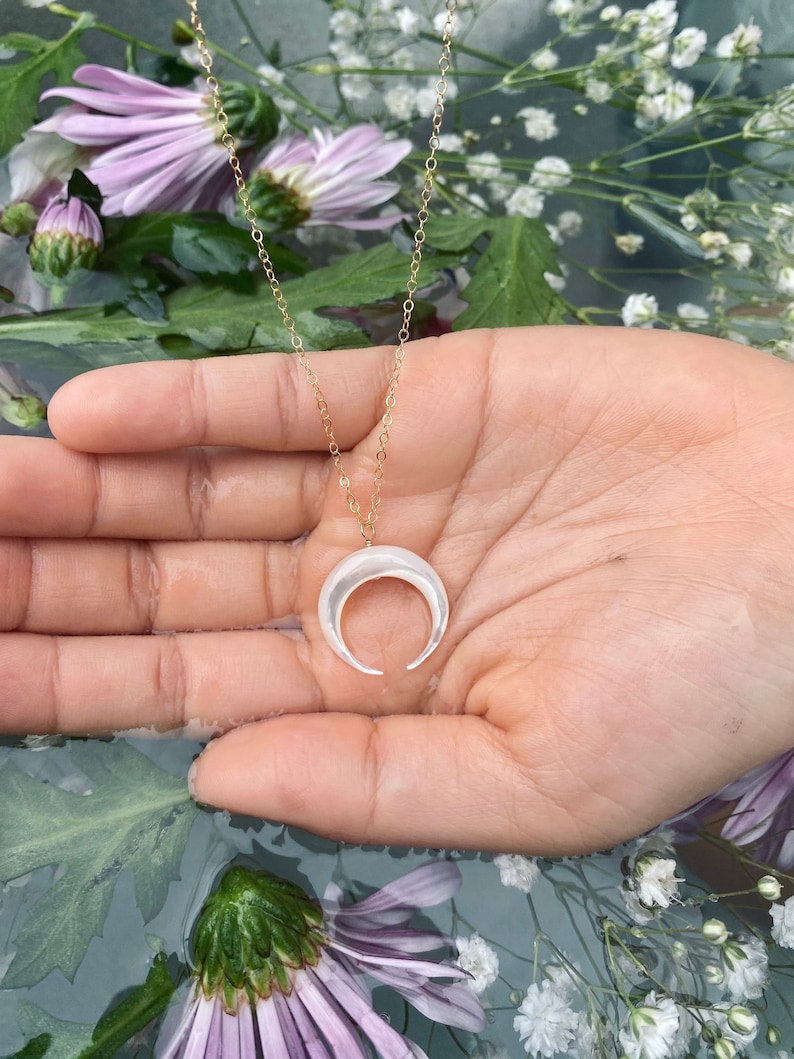 以下が含まれることがあります： 白い三日月型のペンダントが付いた金のチェーンネックレス。ネックレスは花柄の背景に置かれた手に置かれています。