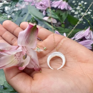 以下が含まれることがあります： 白い三日月型のペンダントが付いたゴールドチェーンネックレス。ペンダントは真珠母貝製で、繊細で女性らしいデザインです。