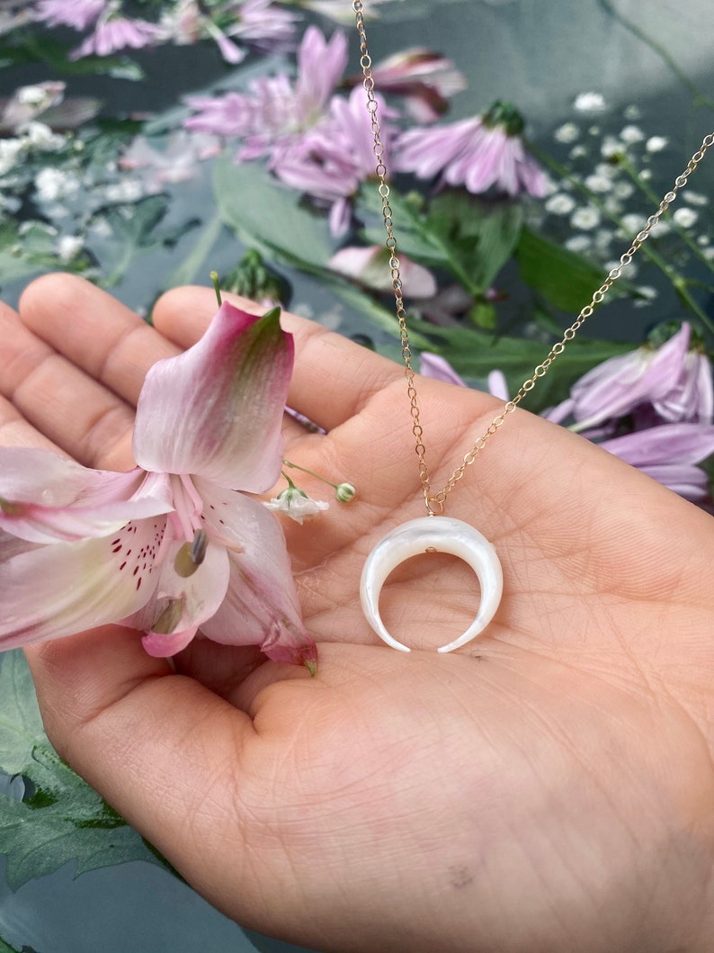 以下が含まれることがあります： 白い三日月型のペンダントが付いたゴールドチェーンネックレス。ペンダントは真珠母貝製で、繊細で女性らしいデザインです。