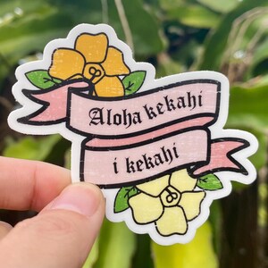 May include: A pink and white sticker with yellow flowers and the text "Aloha kekahī i kekahī".