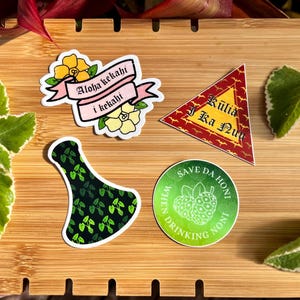 May include: Four stickers on a wooden surface. The stickers are all different designs. One sticker is pink and white with the text "Aloha kekahli i kekahli". Another sticker is red and gold with the text "Kuliia i Ka Nui". A third sticker is green and black with a bottle shape. The final sticker is green and white with the text "Save Da Honl When Drinking Noni".