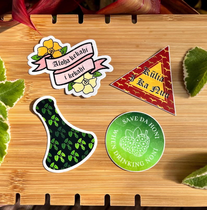May include: Four stickers on a wooden surface. The stickers are all different designs. One sticker is pink and white with the text "Aloha kekahli i kekahli". Another sticker is red and gold with the text "Kuliia i Ka Nui". A third sticker is green and black with a bottle shape. The final sticker is green and white with the text "Save Da Honl When Drinking Noni".