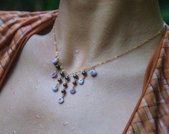 May include: A gold chain necklace with a pendant made of small, dark purple and white beads. The necklace is worn by a person with a brown and white patterned shirt.