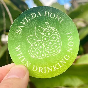 May include: A green and white round sticker with the text "Save Da Honi When Drinking Noni" and a white illustration of a noni fruit.