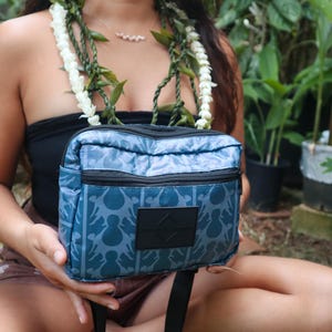 May include: A teal and blue patterned crossbody bag with a black strap and zipper. The bag features a repeating design and a black square patch. The bag is held in front of a person wearing a black top and a flower lei.