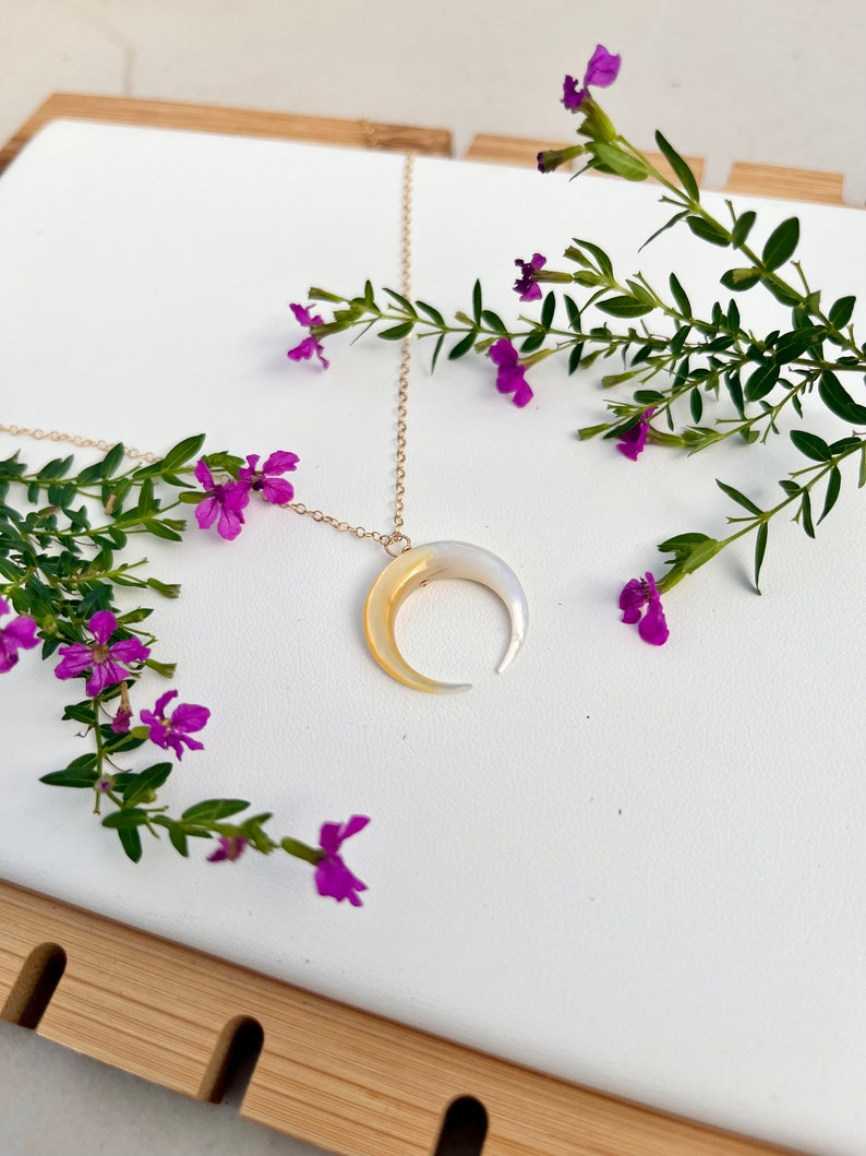 以下が含まれることがあります： 白い真珠貝と黄色の真珠貝で作られた三日月形のペンダントが付いた金のチェーンネックレス。ネックレスは紫の花が咲いた木製の表面に置かれています。