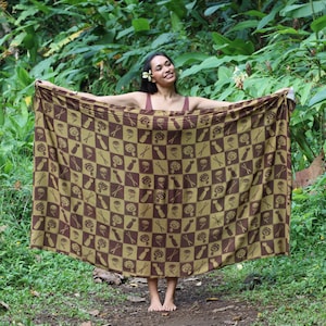 May include: A brown and olive green patterned sarong with a repeating design of stylized flowers and other shapes. The sarong is held open, displaying the full pattern. The background is lush green foliage.