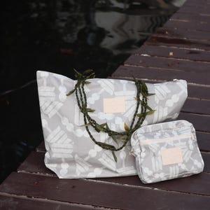 May include: A gray tote bag and matching small pouch with a white pattern. Both bags have a light pink leather patch and a zipper closure. A green garland rests on the bags, which are on a wooden dock.