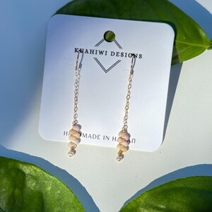 May include: Gold chain drop earrings featuring pale pink spiral-shaped beads and small white pearls at the base. The earrings are attached to a white display card printed with the text KUAHIWI DESIGNS and MADE IN HAWAII.