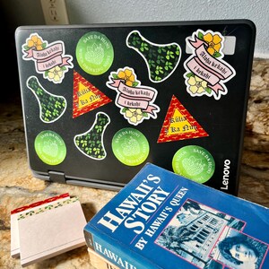 May include: A laptop computer with several stickers on the lid. The stickers are green, pink, and yellow and feature Hawaiian-themed designs and text. The text on the stickers includes "Aloha kekahi i kekahi", "Kūlia Ka Nu", and "Save Da HonI". The laptop is closed and sitting on a table next to a stack of books. The books are blue and have the title "Hawaii's Story" and "Hawaii's Story: The Legends and Myths of Old Hawaii".