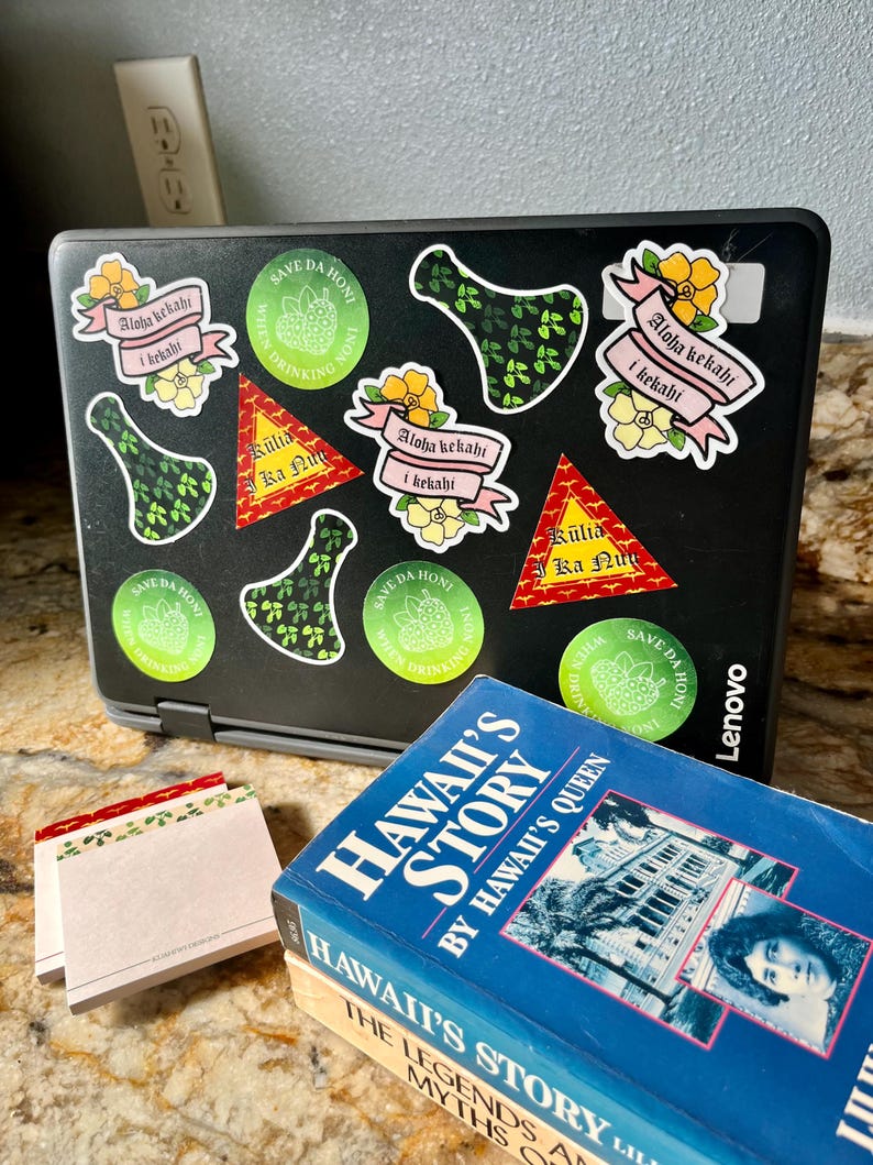 May include: A laptop computer with several stickers on the lid. The stickers are green, pink, and yellow and feature Hawaiian-themed designs and text. The text on the stickers includes "Aloha kekahi i kekahi", "Kūlia Ka Nu", and "Save Da HonI". The laptop is closed and sitting on a table next to a stack of books. The books are blue and have the title "Hawaii's Story" and "Hawaii's Story: The Legends and Myths of Old Hawaii".