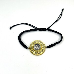 Puede incluir: Una pulsera dorada con un colgante circular con un diseño de moneda de plata. El colgante está rodeado por un patrón geométrico. La pulsera está hecha de cordón negro tejido, con nudos corredizos ajustables.