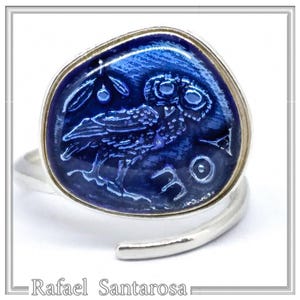 Könnte beinhalten: Ein silberner Ring mit einem blauen Emaille-Eulen-Design. Die Eule ist mit ausgebreiteten Flügeln und einem Zweig mit Oliven dahinter dargestellt. Der Ring ist ein Statement-Piece mit einem einzigartigen Design.