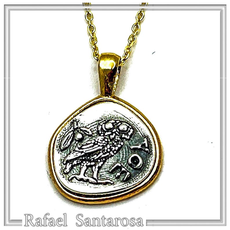 May include: A gold-toned necklace featuring a pendant with a silver-colored, irregular, rounded coin-like design. The pendant depicts an owl in profile, an olive branch, and the letters AOE. The chain is a delicate gold-toned link style.