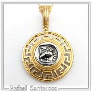 May include: A gold pendant with a round silver coin featuring an owl design. The pendant is surrounded by a gold frame with a Greek key pattern. The pendant is attached to a gold chain.