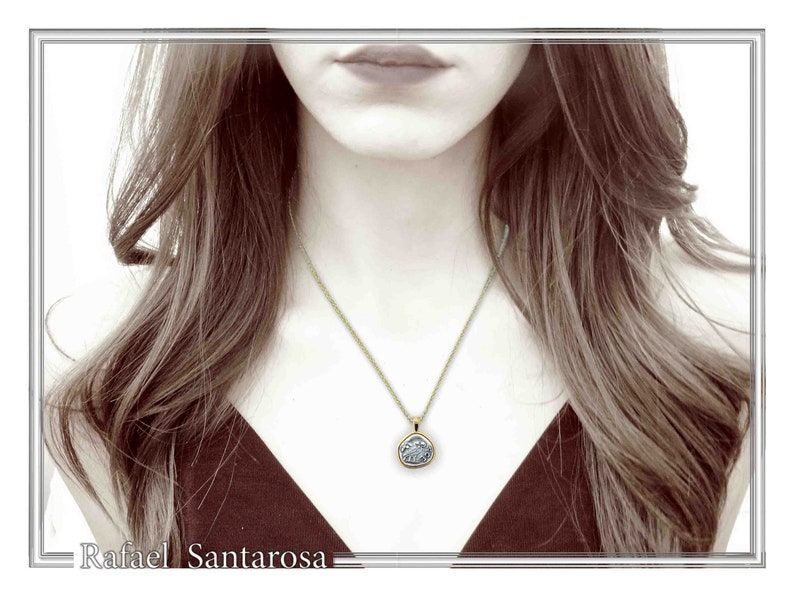 May include: A gold chain necklace featuring a circular pendant with a textured, silver-toned center. The pendant has a smooth gold-colored border and hangs from a delicate gold link chain. The text Rafael Santarosa is visible at the bottom.