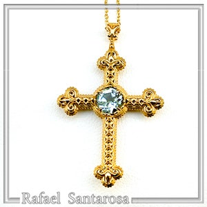 Peut inclure: Un pendentif en forme de croix en or avec une pierre précieuse bleu clair au centre. La croix est ornée de détails complexes et est suspendue à une chaîne délicate.