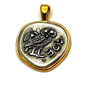 May include: A circular pendant featuring a silver-toned coin design with an owl and an olive branch, encased in a polished gold-toned metal frame. The coin surface displays the Greek letters Alpha, Omicron, and Sigma, with a gold-toned bail at the top.