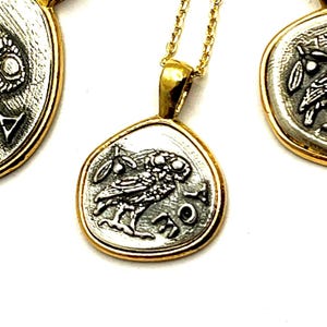 May include: A gold-toned pendant necklace featuring a silver-colored coin charm with an owl and olive branch design. The coin has the letters AOE inscribed on the right side. The pendant hangs from a delicate gold-toned chain, with two similar coin charms visible.