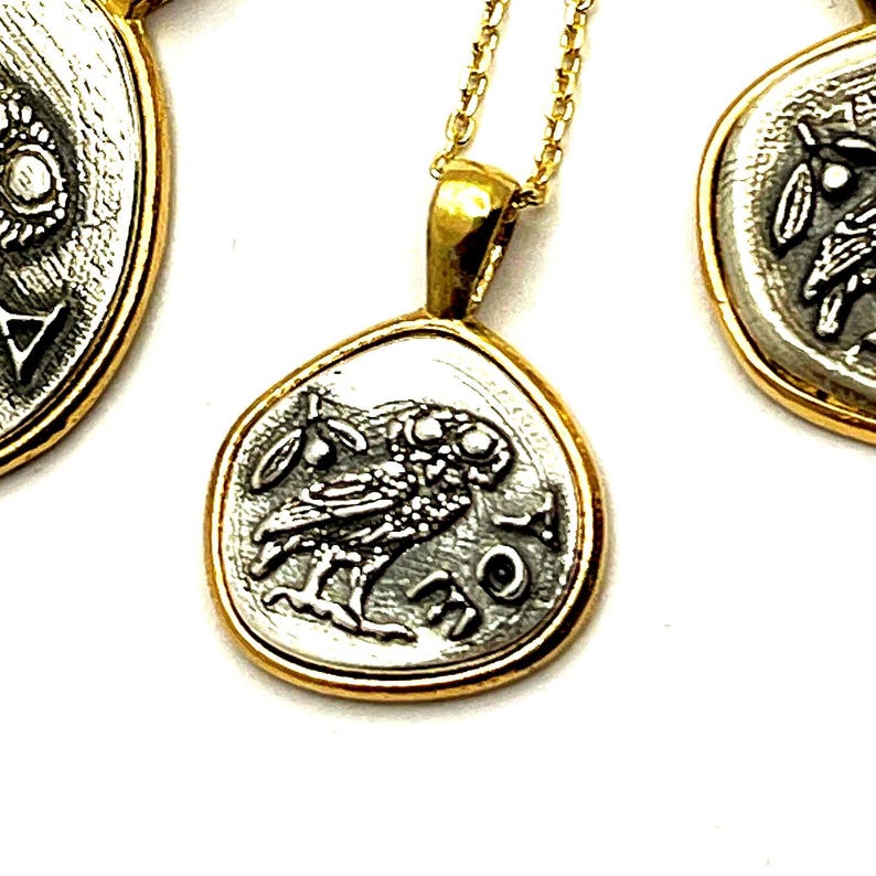 May include: A gold-toned pendant necklace featuring a silver-colored coin charm with an owl and olive branch design. The coin has the letters AOE inscribed on the right side. The pendant hangs from a delicate gold-toned chain, with two similar coin charms visible.
