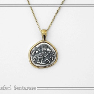 May include: A gold-toned chain necklace featuring a circular pendant with a silver-colored center. The pendant displays an embossed owl figure and the Greek letters AOE. The metal has a textured, aged finish throughout the entire piece.