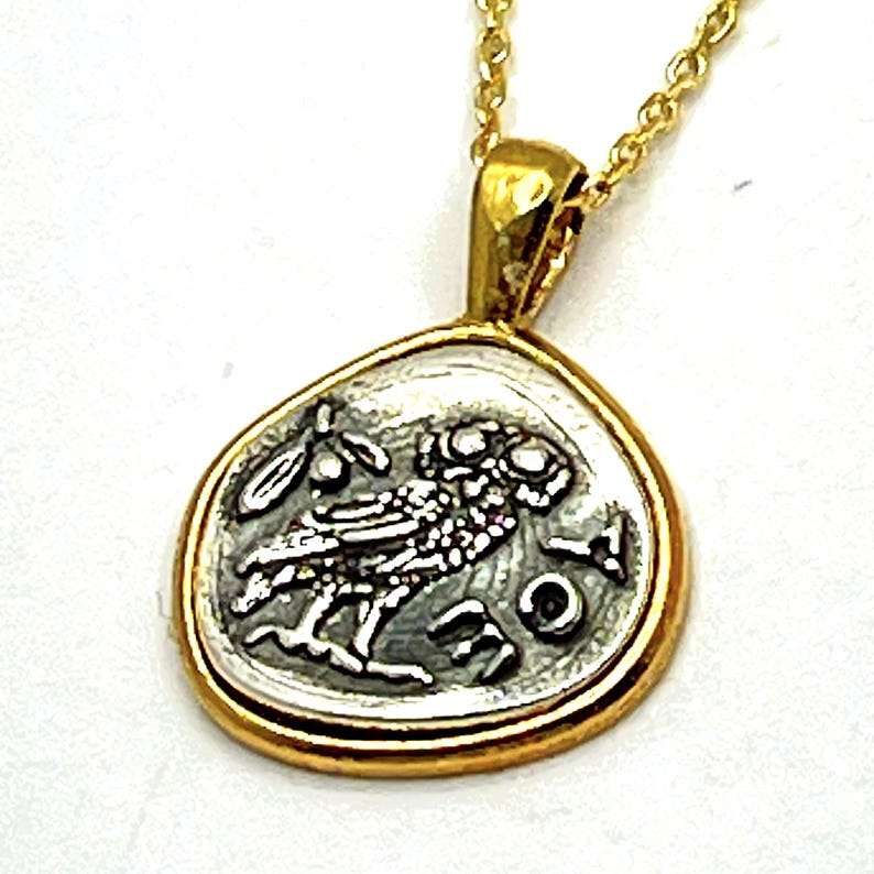 May include: A gold-toned pendant necklace featuring a circular coin-style charm with an irregular edge. The charm displays a silver-colored relief of an owl and an olive branch, accompanied by Greek lettering, suspended from a delicate gold chain.