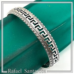 May include: A silver bangle bracelet with a Greek key pattern. The bracelet has a series of connected, angular shapes, with a dotted border. The bracelet is displayed on a green surface. The name "Rafael Santarosa" is at the bottom.