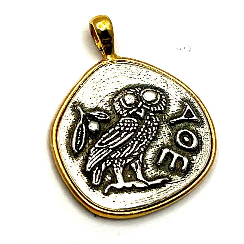May include: A gold-rimmed pendant featuring a silver-toned center with an embossed owl design, an olive branch, and the Greek letters Alpha, Omicron, and Epsilon. The pendant has a smooth, rounded gold bail at the top for attachment to a chain.
