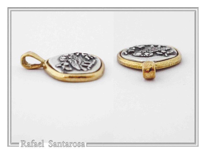 May include: A two-tone pendant featuring a silver-colored oval center with a raised relief design, encased in a smooth gold-colored metal border. The pendant includes a small gold-colored bail at the top for attachment. The text Rafael Santarosa is visible.
