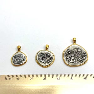 May include: Three silver-toned pendants with gold-toned rims, each featuring an owl and an olive branch design with the letters AOE. A metal ruler is positioned below the pendants, showing the largest piece is approximately 1.5 inches wide.