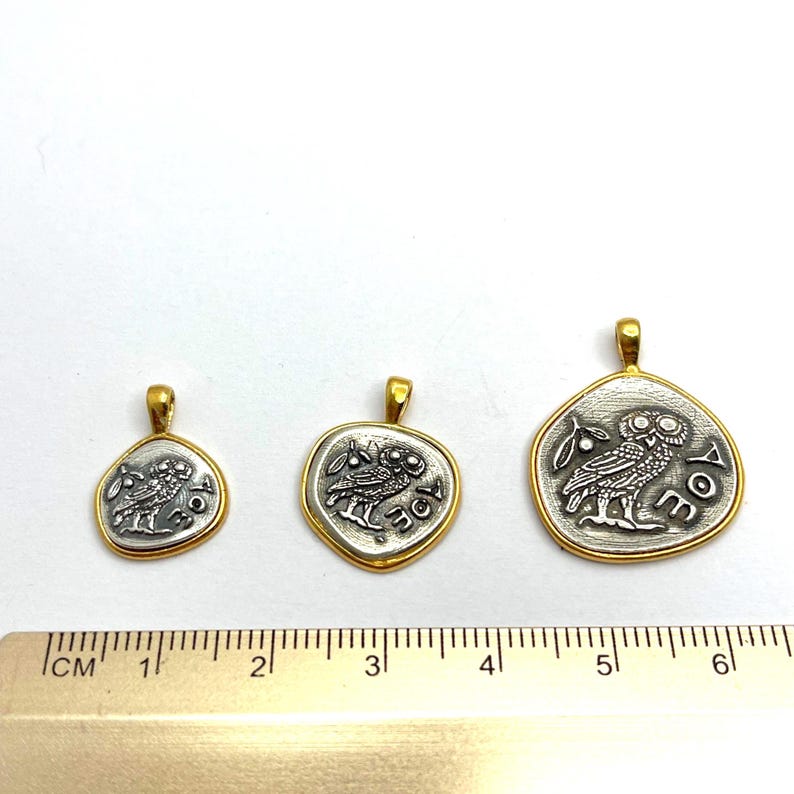 May include: Three silver-toned pendants with gold-toned rims, each featuring an owl and an olive branch design with the letters AOE. A metal ruler is positioned below the pendants, showing the largest piece is approximately 1.5 inches wide.