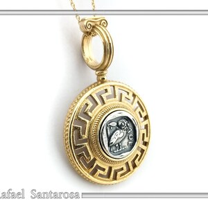 A gold pendant necklace with a round, openwork pendant featuring a Greek key design. The pendant has a smaller round silver coin in the center with an image of an owl and the word 'AΘE'. The pendant is attached to a gold chain.