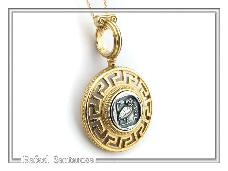 A gold pendant necklace with a round, openwork pendant featuring a Greek key design. The pendant has a smaller round silver coin in the center with an image of an owl and the word 'AΘE'. The pendant is attached to a gold chain.