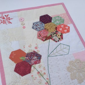 May include: A handmade patchwork quilt with a pink border and a floral design. The quilt features a variety of fabrics, including lace, floral prints, and plain colours. The quilt is stitched together with a variety of colours of thread.