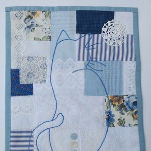 May include: A blue cat outline is stitched onto a patchwork quilt made from white and blue fabric scraps. The quilt is made of various textures, including lace, stripes, and floral patterns.