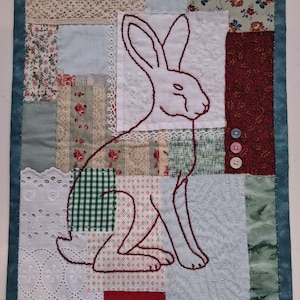 May include: A rectangular fabric wall hanging featuring a patchwork design of various floral, chequered, and solid-coloured textile squares. A dark red embroidered outline of a rabbit sits centrally, accented by three small buttons and lace trim.