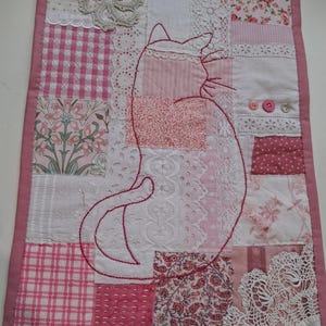 May include: A handmade patchwork quilt with a cat design. The quilt combines pink and white fabrics, such as floral patterns, gingham, and lace. A red embroidered cat outline is featured, and the quilt is finished with a pink border.