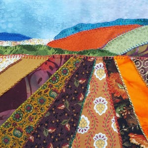 May include: A patchwork quilt featuring a landscape design. The quilt is made from a variety of fabrics in shades of brown, orange, green and blue. The quilt is stitched together with a decorative stitch.