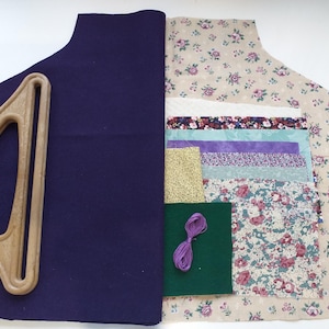 May include: A craft kit featuring a dark blue felt base and a wooden handle. The kit includes various fabric pieces with floral patterns, solid colours, and gold glitter, along with purple embroidery thread. The kit is designed for creating a bag.
