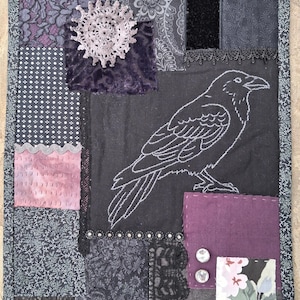May include: A rectangular fabric wall hanging featuring a patchwork design of black, grey, and purple textiles. The piece includes an embroidered white raven, a circular lace doily, black lace trim, two silver buttons, and a floral fabric square.