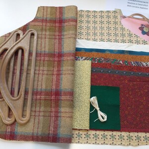 May include: A sewing kit featuring a selection of fabrics, wooden handles, and a small pink bag with a floral design. The fabrics display patterns like plaid and floral, alongside solid colours. White thread and green felt are also present.