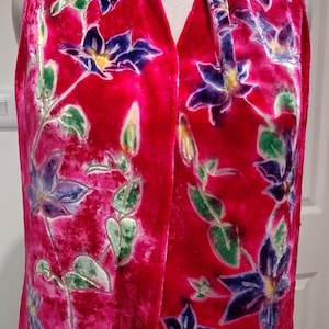 May include: A bright pink scarf with a floral design. The scarf displays a pattern of purple and blue flowers with green leaves and stems. The fabric has a soft, velvety texture. The scarf is draped over a mannequin.