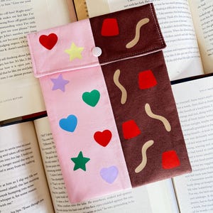 May include: A two-tone pink and brown fabric pouch with a white button fastening. The pink side has colourful felt heart, star, and other shapes. The brown side has felt shapes resembling chocolate and sprinkles. The pouch is open on a book.