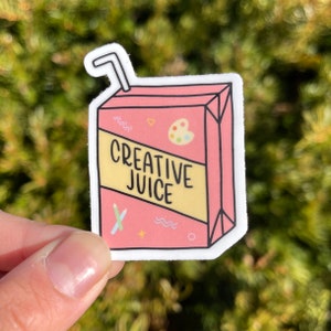 May include: A pink and yellow sticker with a cartoon illustration of a juice box. The juice box has a straw sticking out of the top and says "Creative Juice" on the front. The juice box is decorated with colorful paintbrushes, a paint palette, and other artistic elements.