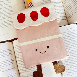 May include: A whimsical bookmark shaped like a pink cake with three red cherry-like decorations and brown felt feet. The bookmark has a smiling face and a white frosting layer. It is placed on open books.