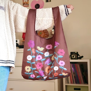 May include: A brown reusable shopping bag with a floral print. The flowers are in shades of pink, purple, and yellow. The bag is folded and ready to be used.