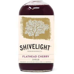 May include: A glass bottle of Shivelight Flathead Cherry Shrub, a premium beverage. The bottle is filled with a dark red liquid and has a black cap. The label features a stylized tree design and the text "Shivelight Premium Beverage Co. Flathead Cherry Shrub Makes 16 Cocktails or 12 Sodas".