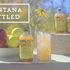 May include: A glass of iced tea with a straw, a bottle of Shivelight Bitteroot Apple & Cinnamon Syrup, and a bowl of red and green apples on a white wooden table. The text "MONTANA BOTTLED" is in a white rectangle on a green background.